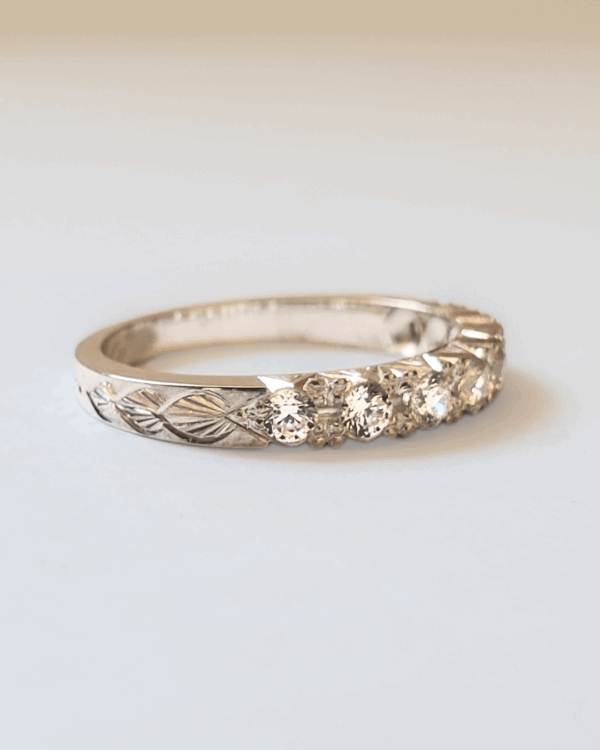 Bague de fiançailles ou alliance en or gris pavée de diamants et gravée de motif végétal. Posée sur fond clair.
