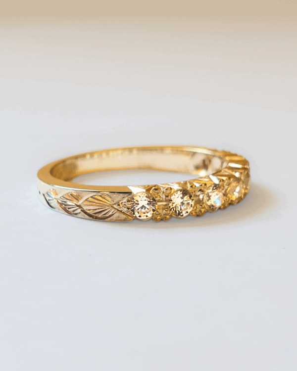 Bague de fiançailles ou alliance en or jaune pavée de diamants et gravée de motif végétal. Posée sur fond clair.