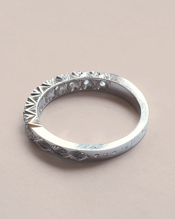 Bague de fiançailles ou alliance en or gris gravée de motif végétal, pavée de diamants au sertissage relevé de gravures sur la tranche du bijou. Posée sur fond clair.