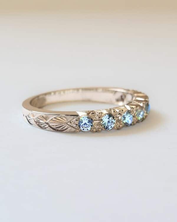 Bague de fiançailles ou alliance en or gris pavée de topazes et gravée de motif végétal. Posée sur fond clair.
